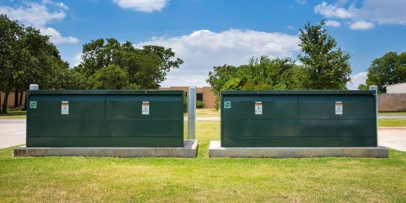 Two green underground distribution switchgear boxes on concrete pads. In the foreground is green grass. In the background are trees and a blue sky.